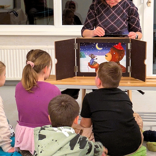 Mehrere Kinder sitzen auf Matten und Kissen vor einem Kamishibai-Erzähltheater. Eine erwachsene Person zeigt und liest eine Geschichte mit bunten Bildern.