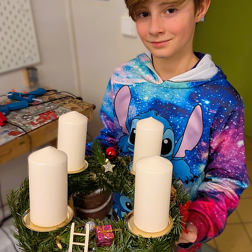 Ein Junge hält einen selbstgebastelten Adventskranz mit vier weißen Kerzen, kleinen Geschenken und Miniaturleiter.