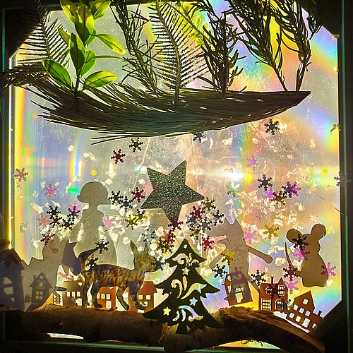 Ein weihnachtlich dekoriertes Fensterbild mit Scherenschnittfiguren und Naturmaterialien wird von Licht durchleuchtet und erzeugt bunte Lichtreflexe.