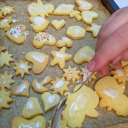 Ein Kinderhand verziert frisch gebackene Plätzchen auf einem Backblech mit weißem Zuckerguss und bunten Streuseln.