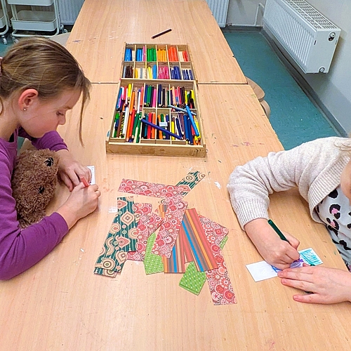 Zwei Mädchen sitzen an einem Tisch und gestalten bunte Lesezeichen mit Buntstiften und Musterpapier. Eines der Kinder hält dabei ein Kuscheltier.