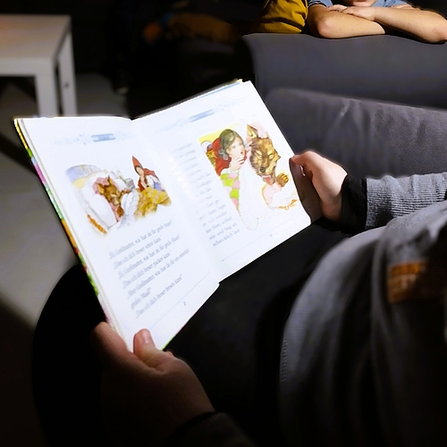 Ein Kind liegt auf einem Sofa und hört gespannt zu, wie jemand bei gedämpftem Licht aus einem Märchenbuch vorliest.