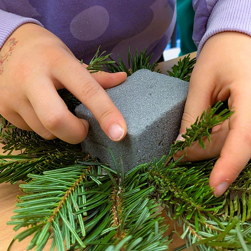 Zwei Kinderhände stecken Tannenzweige in einen grauen Steckschwamm, um ein Adventsgesteck zu gestalten.