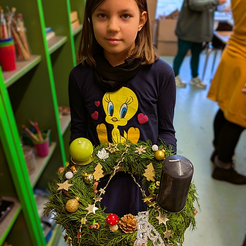 Ein Mädchen präsentiert ihren Adventskranz mit einer schwarzen und einer grünen Kerze, geschmückt mit Sternen, Pilzen und goldenen Kugeln.