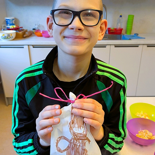Ein Junge mit Brille hält stolz eine weiße Tüte mit einer Zeichnung hoch, im Hintergrund ist die Arbeitsfläche der Küche.