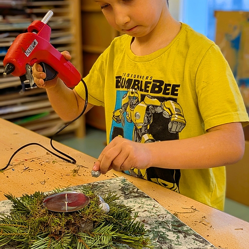 Ein Junge arbeitet konzentriert an einem kleinen Adventsgesteck mit einer roten Kerze, während er eine Heißklebepistole in der Hand hält.