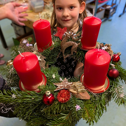 Ein Kind hält einen opulent geschmückten Adventskranz mit vier großen roten Kerzen und vielen gold-roten Deko-Elementen.